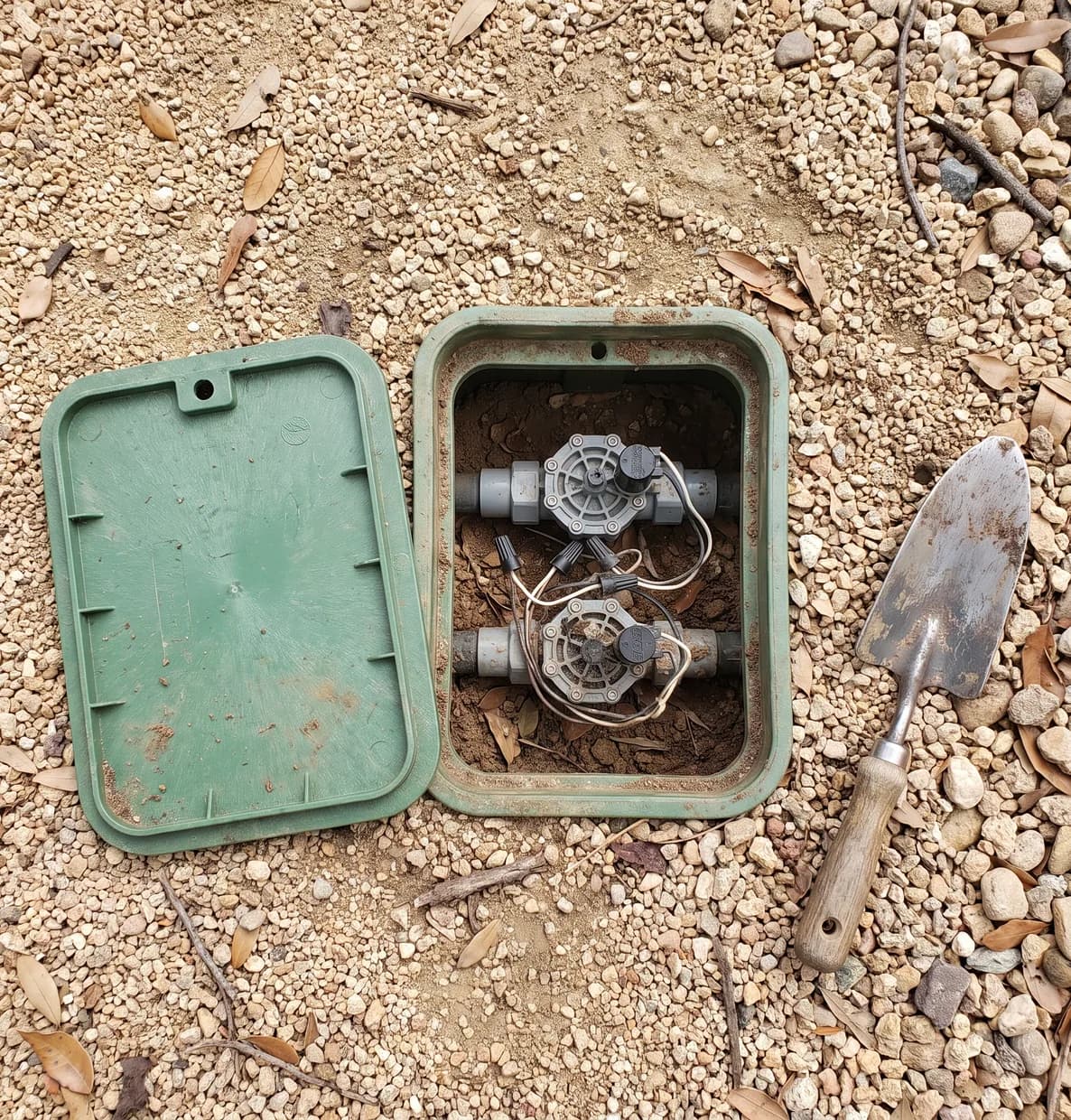 Open irrigation valve box in gravel with two valves and solenoids, the green lid set aside and a trowel nearby