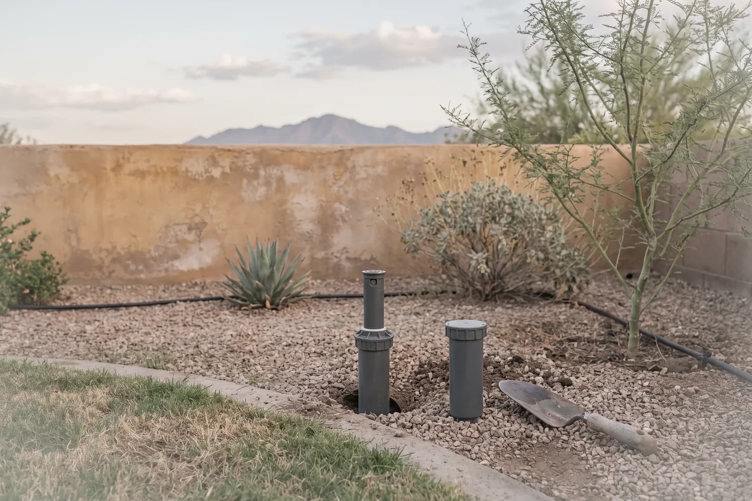 Two pop-up sprinkler heads at a gravel and turf edge, one lifted from its opening and one new spare beside a trowel