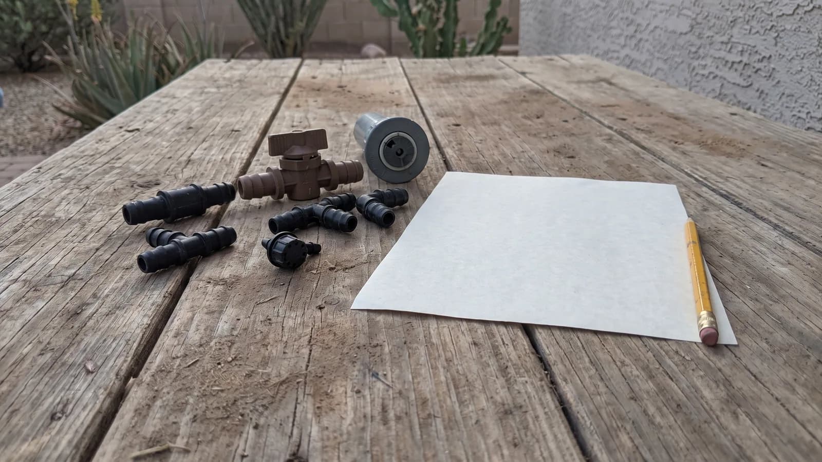Irrigation fittings, a drip emitter, a valve and a pop-up sprinkler head laid out on a patio worktable beside a blank sheet of paper and a pencil